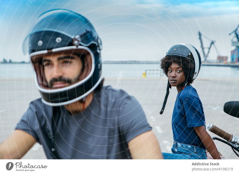 Positive young multiracial couple in helmets sitting near motorbike at seaside biker motorcycle promenade style together date relationship ride trip rest
