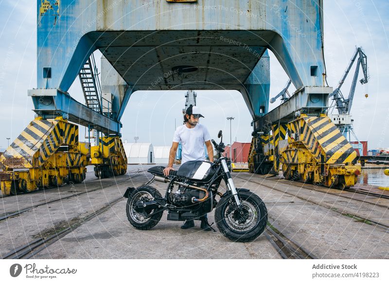 Cal young ethnic guy with motorbike near cranes working near port man biker motorcycle industrial serious brutal transport seaside confident male casual cap
