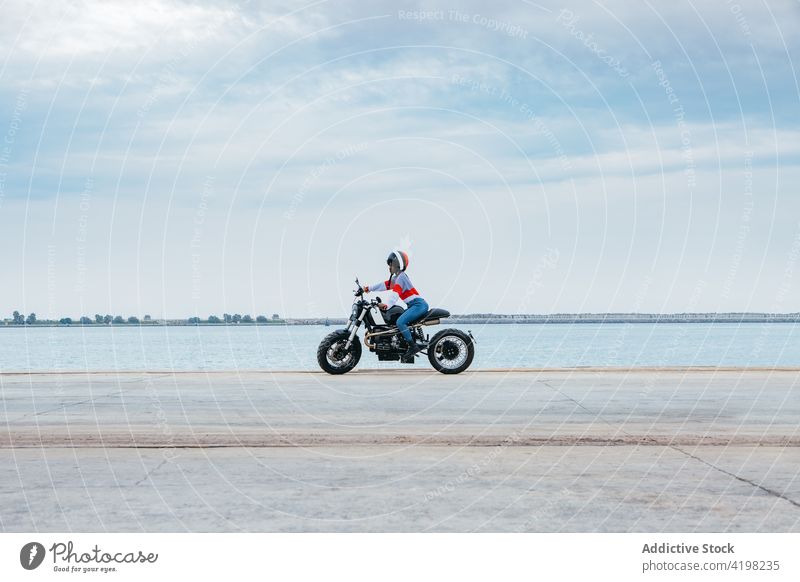 Anonymous trendy lady riding motorbike near sea woman ride motorcycle road style alone biker trip enjoy travel vehicle female young cool helmet casual