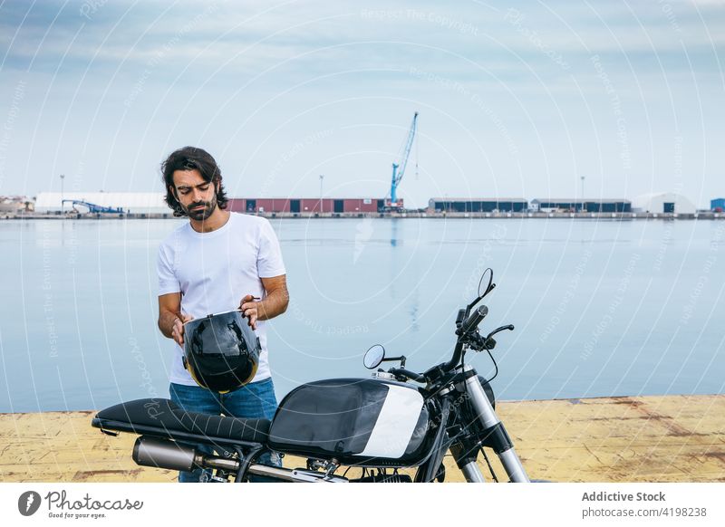 Serious young ethnic man in a motorbike on embankment helmet motorcycle seaside confident brutal serious style biker casual male t shirt jeans beard transport