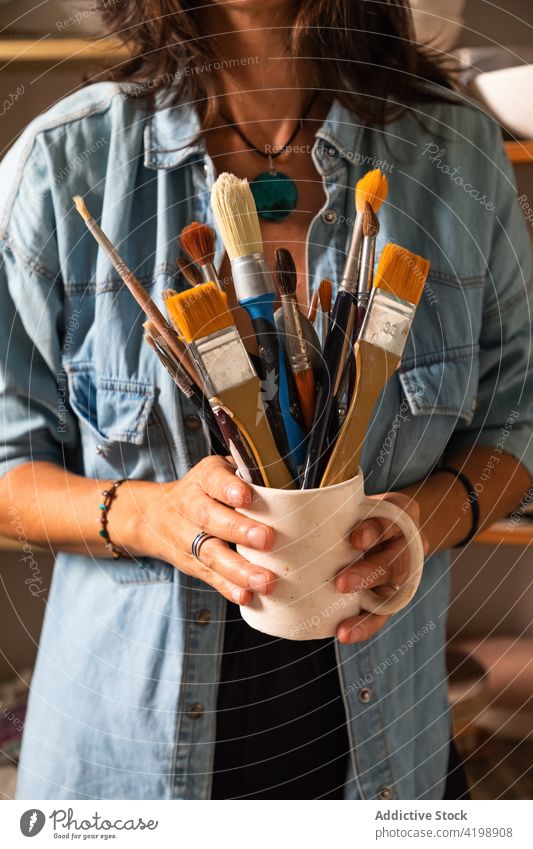 Anonymous craftswoman holding cup with assorted brushes in workshop paintbrush master pottery clayware cheerful artisan various tool create female adult casual