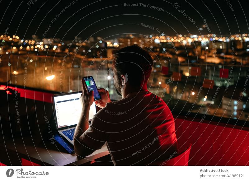 Focused man using smartphone and laptop during online work on rooftop at night message remote evening freelance concentrate communicate connection male young