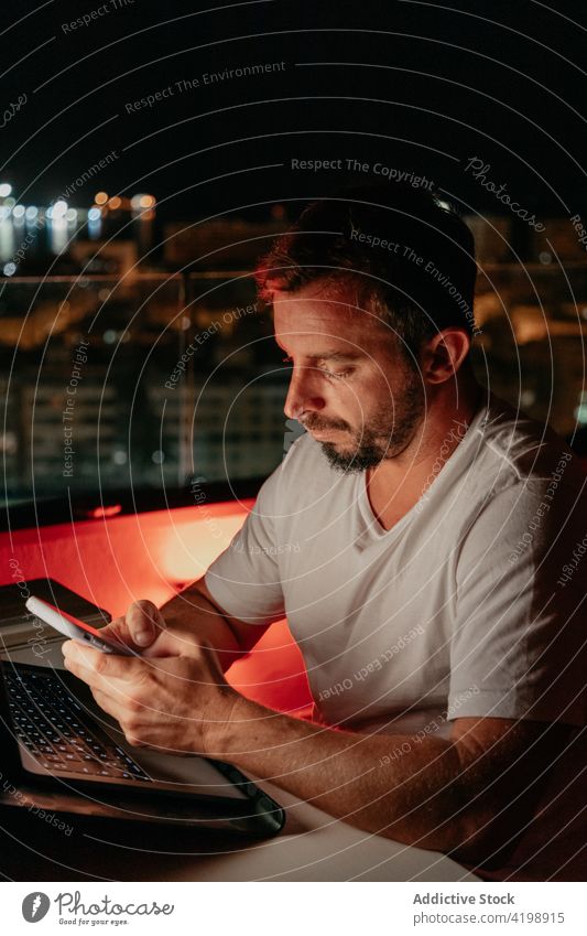 Focused man using smartphone and laptop during online work on rooftop at night message remote evening freelance concentrate communicate connection male young