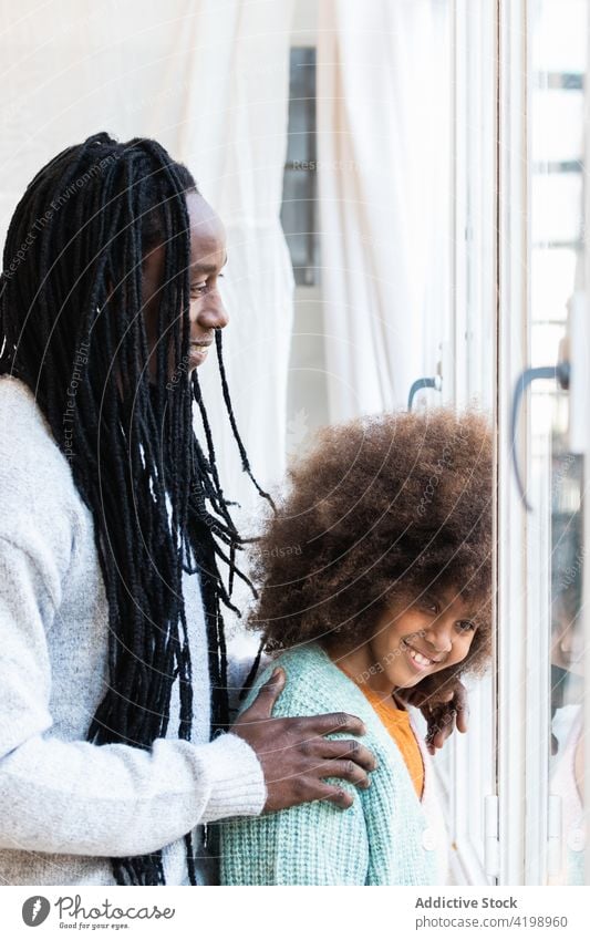 Black father with daughter standing near window close cheerful carefree bonding positive tender smile hug girl man dad childhood kid afro parent casual love