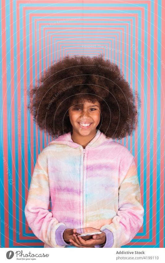 Delighted ethnic girl with smartphone standing in studio afro hairstyle smile cheerful teenage colorful vivid black african american wall pink blue geometry