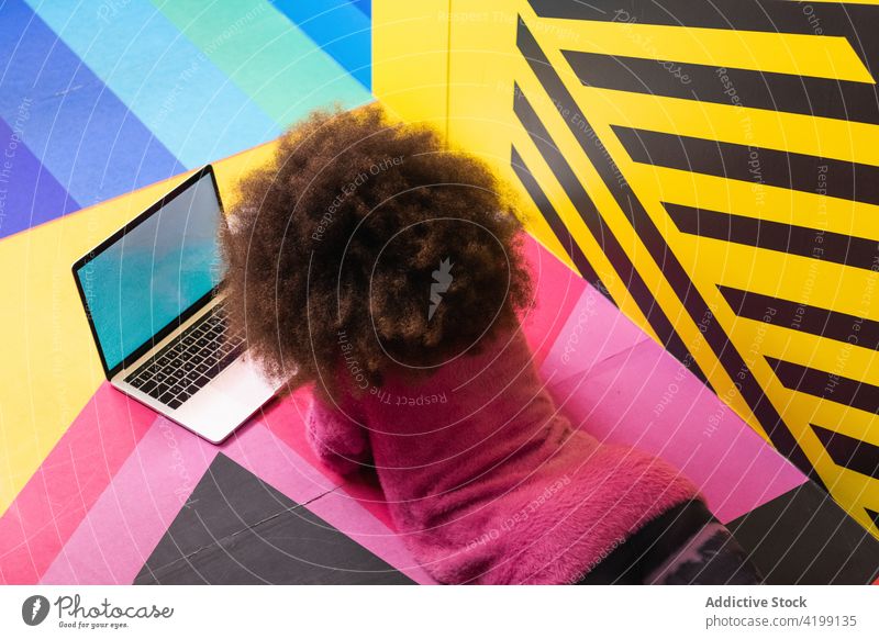 Anonymous black girl using laptop in studio browsing teenage colorful vivid internet social media ethnic african american entertain lying floor netbook surfing