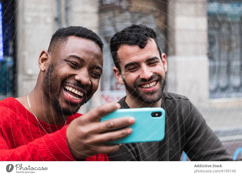 Cheerful diverse friends taking selfie on smartphone on city bench happy moment memory street using gadget device self portrait macho touch chin masculine