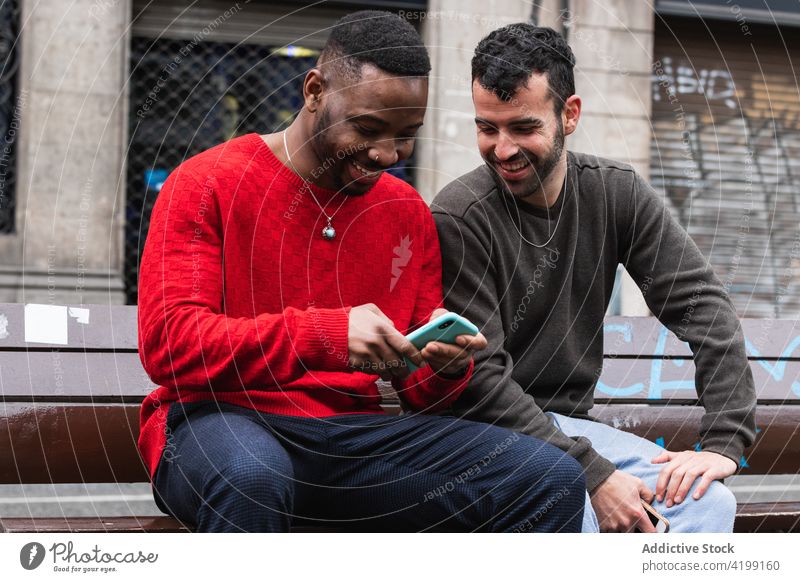Cheerful diverse using smartphone on city bench friend happy street men gadget device macho masculine casual wear together cellphone urban road smile cheerful