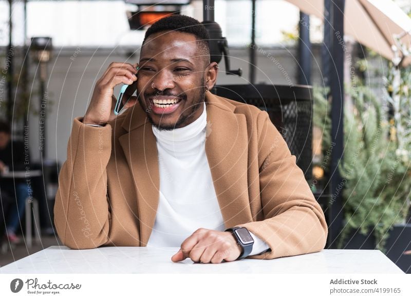 Content black man speaking on smartphone in cafe happy fashion masculine well dressed enjoy table using gadget device communicate cellphone hole fence macho