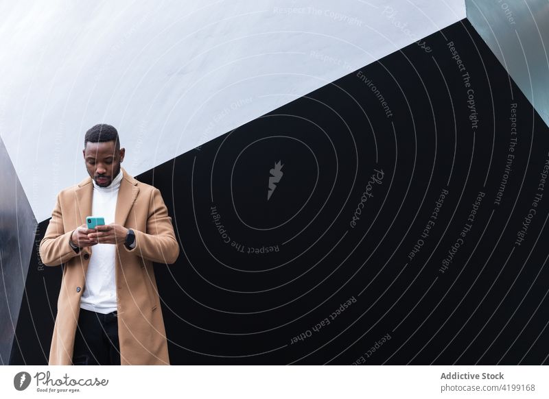 Well dressed black man chatting on smartphone near wall internet online attentive masculine macho using gadget device african american ethnic coat modern