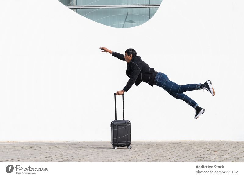 Ethnic tourist with suitcase jumping in air above urban pavement traveler hurry rush fly arm raised stylish man mouth opened building wall gumshoe jeans jacket