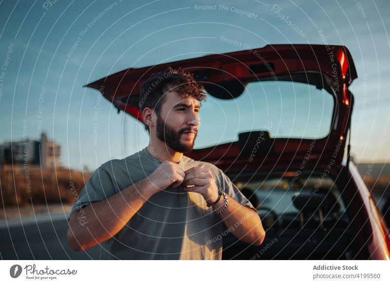 Serious bearded man in car trunk putting clothes on vehicle transport automobile road trip taillight concentrate button male parked putt on t-shirt open alone