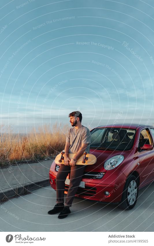 Young bearded skater leaning on car hood and looking away in nature man lean on freedom countryside rural remote road asphalt skateboard thoughtful parked hobby