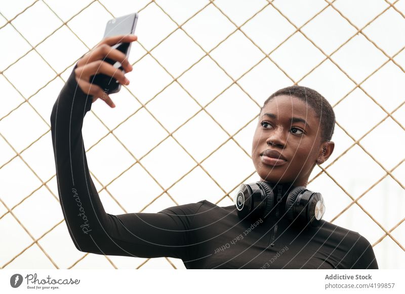 Charming black woman with headphones taking selfie on smartphone self portrait memory moment charming fence using gadget gentle feminine tender device grid sky