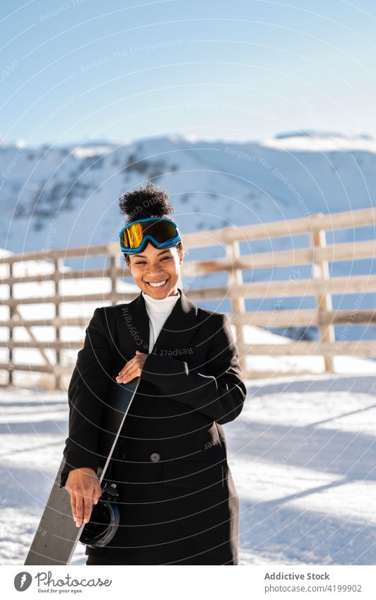 Content black athlete with snowboard in snowy mountains resort style happy nature wintertime woman highland fence weekend sportswoman stylish smile glasses