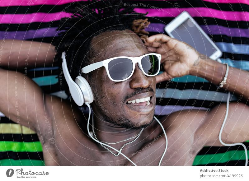 Cheerful black man listening to music in headphones towel summer lawn cheerful song enjoy male african american ethnic carefree lying relax device young