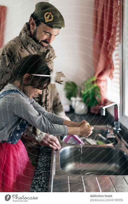 Smiling military father and cute girl washing dishes together daughter soldier kitchen home sink counter man male homecoming arrive serviceman love child