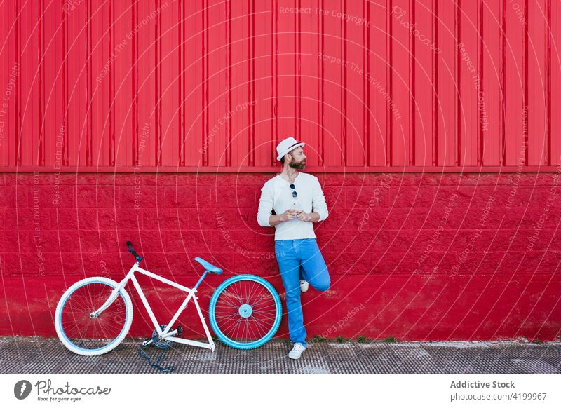Man on smartphone standing near bicycle against red building man relax street trendy style red wall rest fashion thoughtful city sunny urban male beard cool
