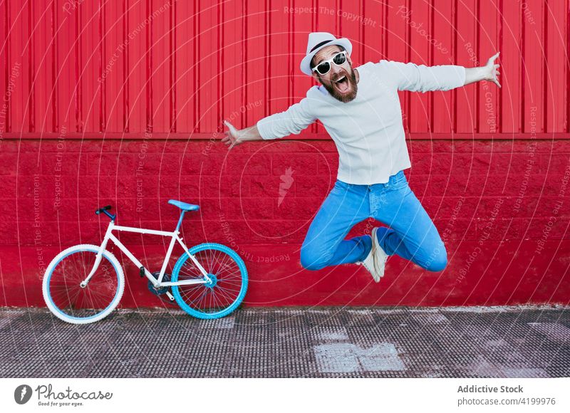 Hipster man jumping near bicycle on city street ride bike cyclist trendy building male beard outfit cap hipster urban modern transport vehicle activity hobby