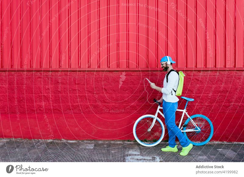 Man on smartphone and headphones walking with bicycle on red building man street trendy style red wall rest fashion city urban male beard cool outfit sunglasses