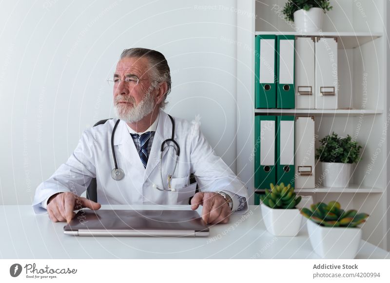 Senior physician in uniform at table in clinic tablet profession man portrait contemplate device gadget online consultant professional stethoscope pensive