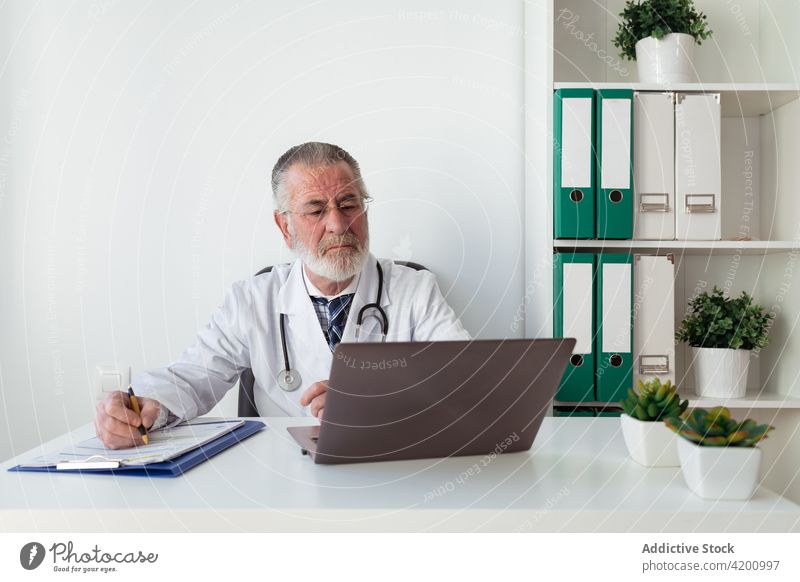 Elderly physician taking notes during video call on laptop in clinic write health care consultation internet remote man using gadget device workplace take note