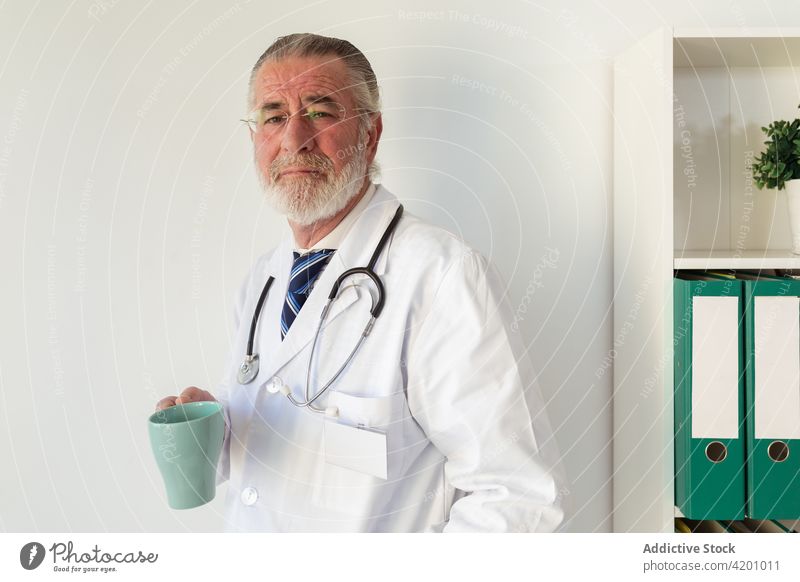 Elderly physician with cup of coffee in clinic doctor uniform professional specialist stethoscope man portrait organize hot drink beverage tea eyeglasses