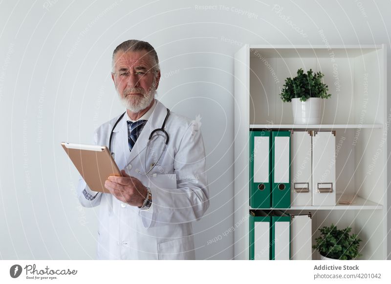 Senior physician in uniform with table in clinic tablet profession man portrait contemplate device gadget online consultant professional stethoscope pensive