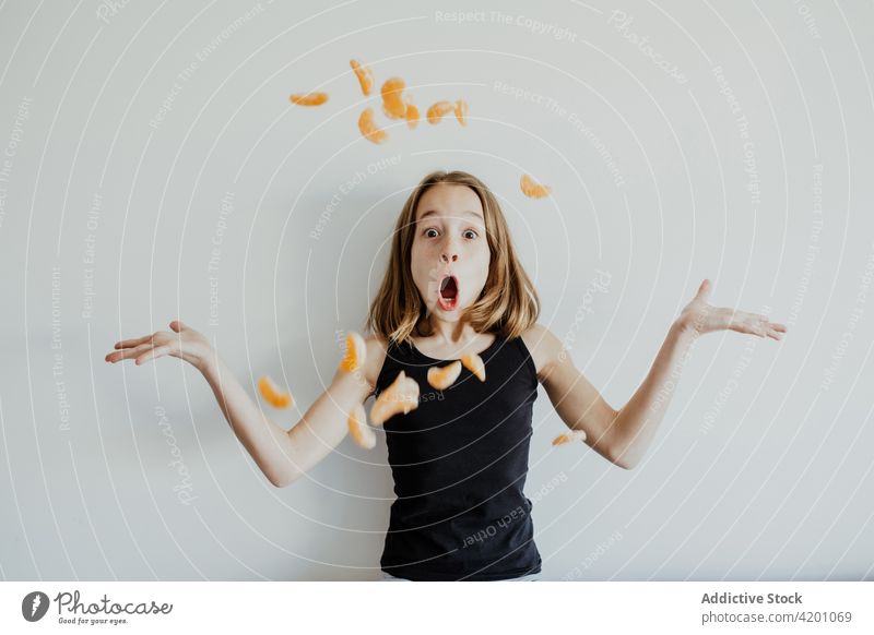 Excited girl throwing tangerine slices and making grimace against white wall child mandarin mouth opened amazed shock having fun shrug excited portrait joy