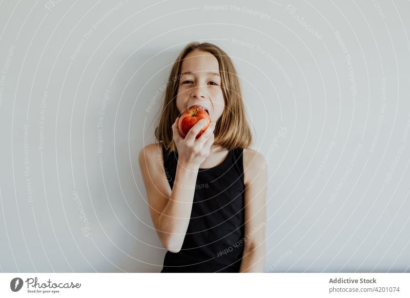 Delighted kid biting apple against white wall girl bite cheerful smile eat fresh fruit vitamin enjoy portrait meal happy ripe child preteen casual top