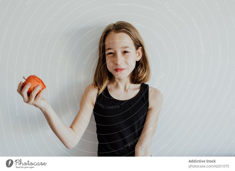 Delighted kid biting apple against white wall girl bite cheerful smile eat fresh fruit vitamin enjoy portrait meal happy ripe child preteen casual top