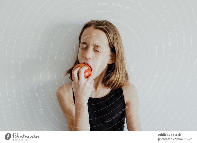Delighted kid biting apple against white wall girl bite eat fresh fruit vitamin enjoy portrait meal ripe child preteen casual top human face daylight delicious