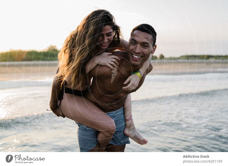 Cheerful black man giving girlfriend piggyback ride against sea couple relationship having fun honeymoon ocean trip vacation happy wavy boyfriend love smile