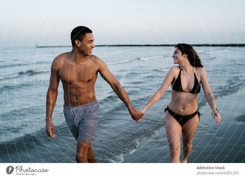 Glad diverse couple holding hands while walking on sea beach relationship love romantic tourism vacation glad honeymoon abdomen shorts belly body six pack coast