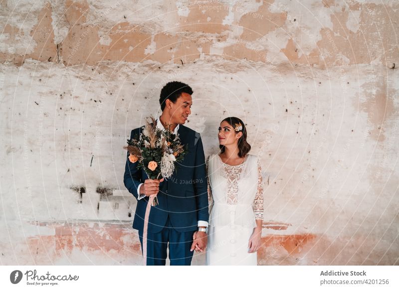 Sincere multiethnic newlywed couple holding hands near rough wall wedding vintage relationship love bouquet smile celebrate botany aroma bloom flower aged