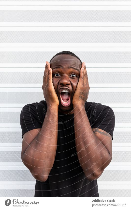 Amazed black man screaming near striped wall touch face amazed yell astonish surprised omg overwhelmed portrait mouth opened t shirt shout shock ethnic
