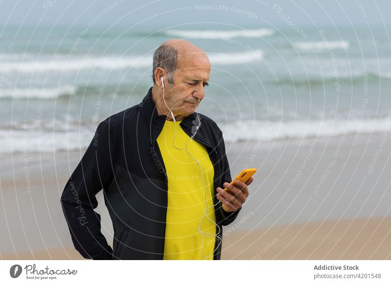 Elderly sportsman listening to music near sea shore smartphone using wave training break male fitness elderly aged senior earphones athlete healthy coast ocean