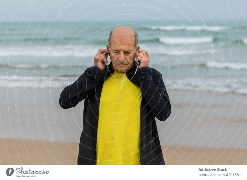 Senior sportsman putting on earphones on seashore put on training fitness beach listen music male athlete workout aged elderly senior lifestyle vitality