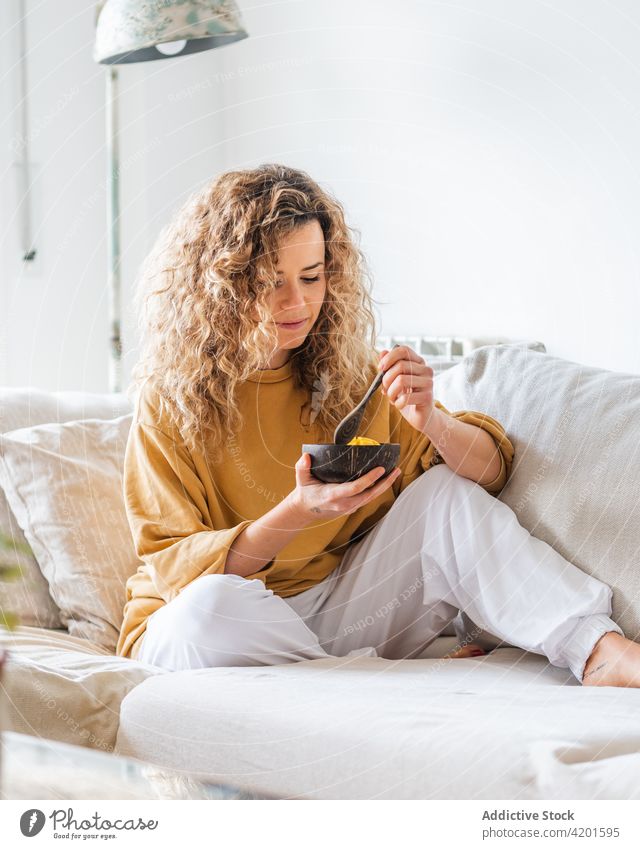Blonde female sitting with bowl of healthy food on sofa woman positive apartment curly hair mango young room home eat style trendy tasty living room indoors