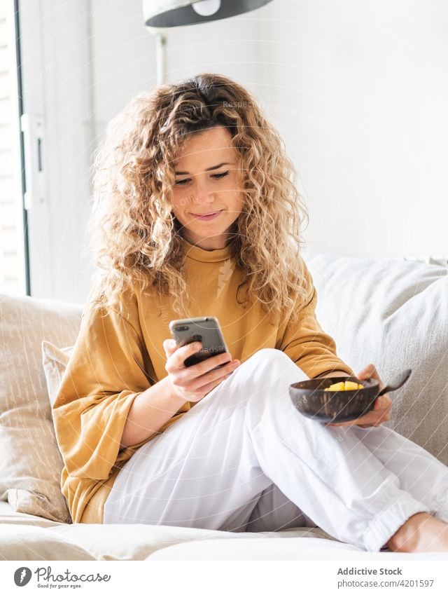 Cheerful female browsing smartphone while sitting with bowl of food on sofa woman positive cheerful using text message apartment smile surfing mobile curly hair