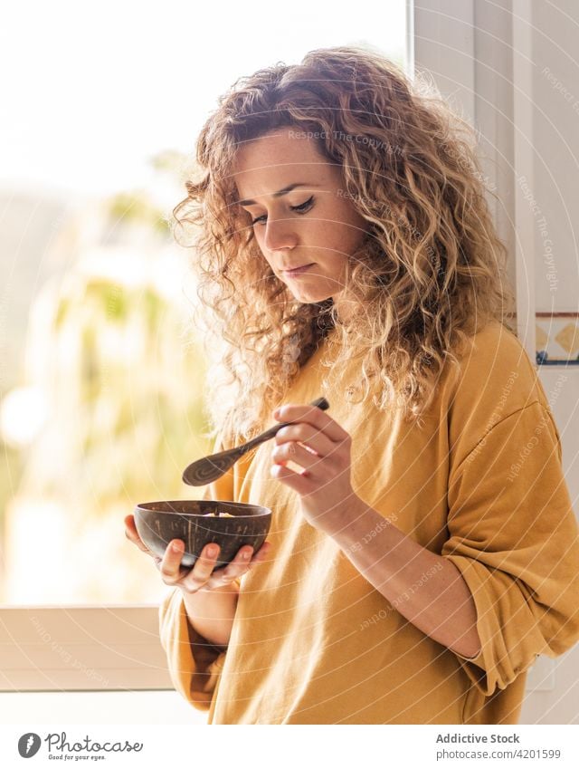 Calm woman eating healthy breakfast in apartment calm pensive food thoughtful room bowl female curly hair tranquil charming spoon wooden window home light lady
