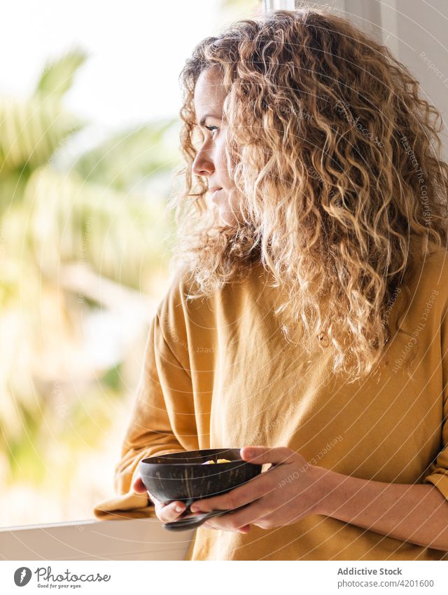Calm woman eating healthy breakfast in apartment calm pensive food thoughtful room bowl female curly hair tranquil charming spoon wooden window home light lady