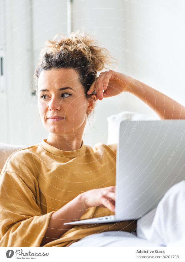 Smiling female sitting with laptop in sofa in light room woman using browsing apartment home device freelance thoughtful positive casual looking away netbook