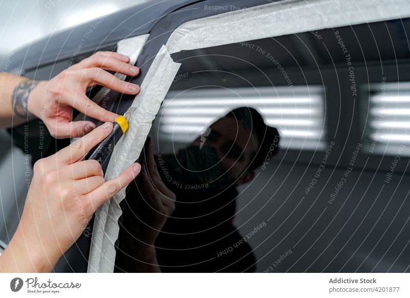 Male mechanic sticking tape on car in repair service man paint prepare workshop auto stationery knife vehicle mask busy professional reflection window