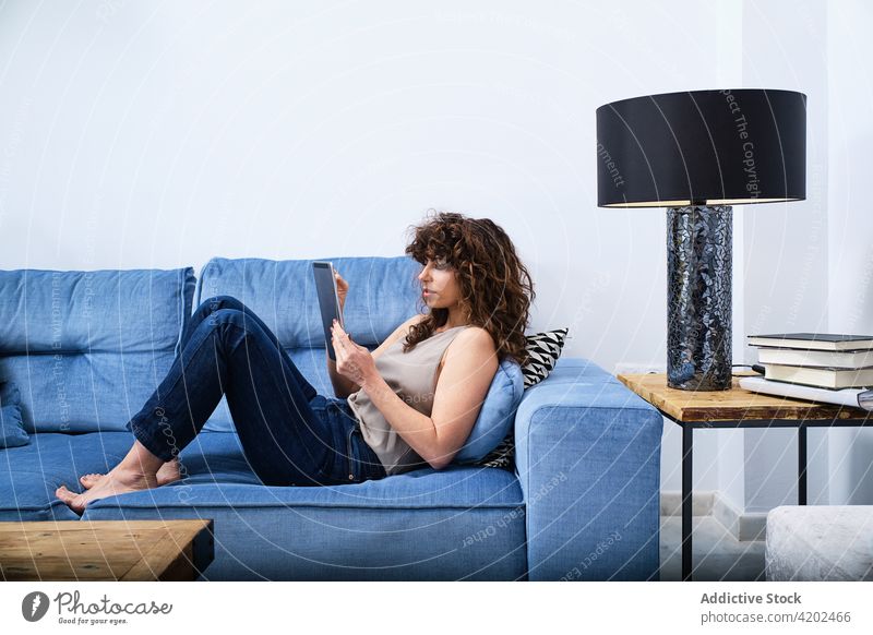 Young woman using tablet while having rest on couch lying browsing sofa room surfing internet relax free time female casual living room chill home comfort
