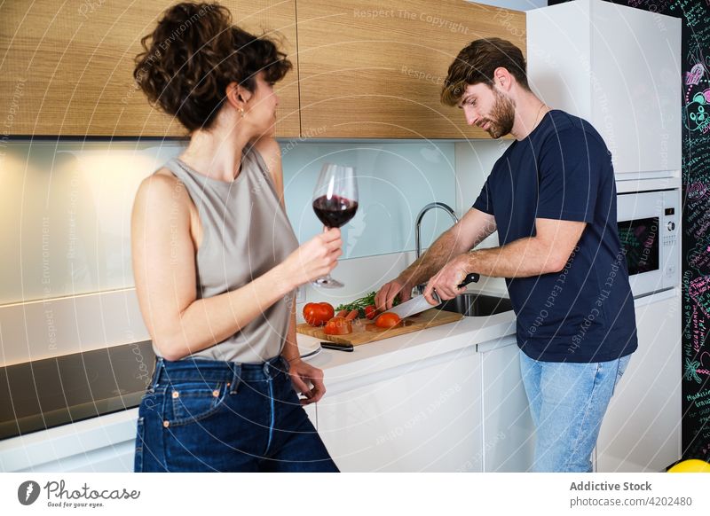 Couple in kitchen with wineglass while cutting vegetables couple cook tomato drink friend counter lifestyle young cabinet home red stove knife sink tap alcohol
