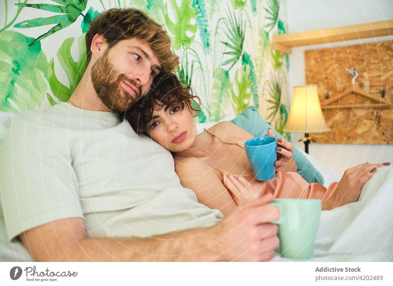 Couple lying in bed together with cups of drink couple home girlfriend boyfriend morning happy young sleepwear light apartment relationship love coffee enjoy