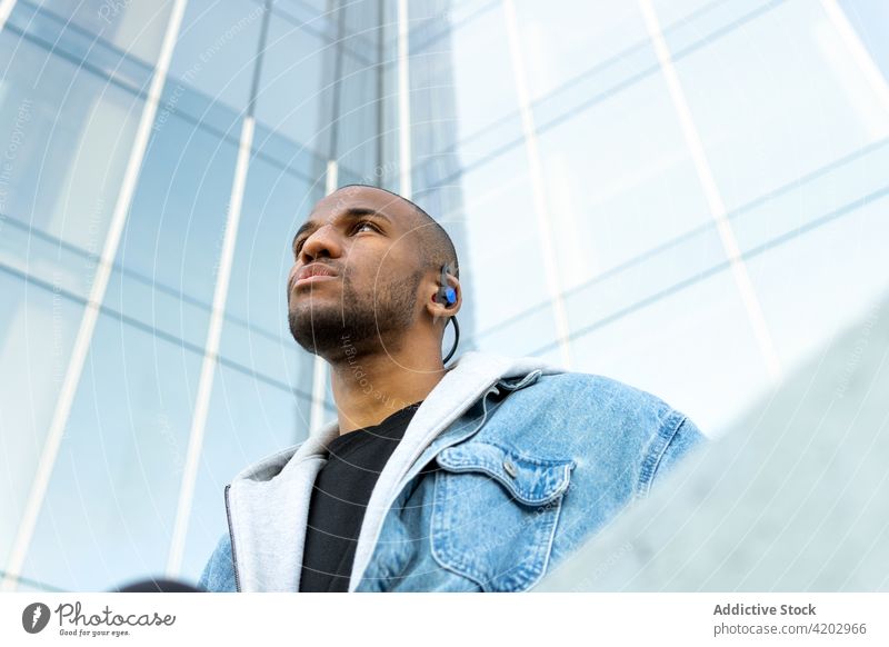 Black man listening to music from earphones in city wireless song dreamy masculine town portrait using device macho brutal building modern street enjoy jacket