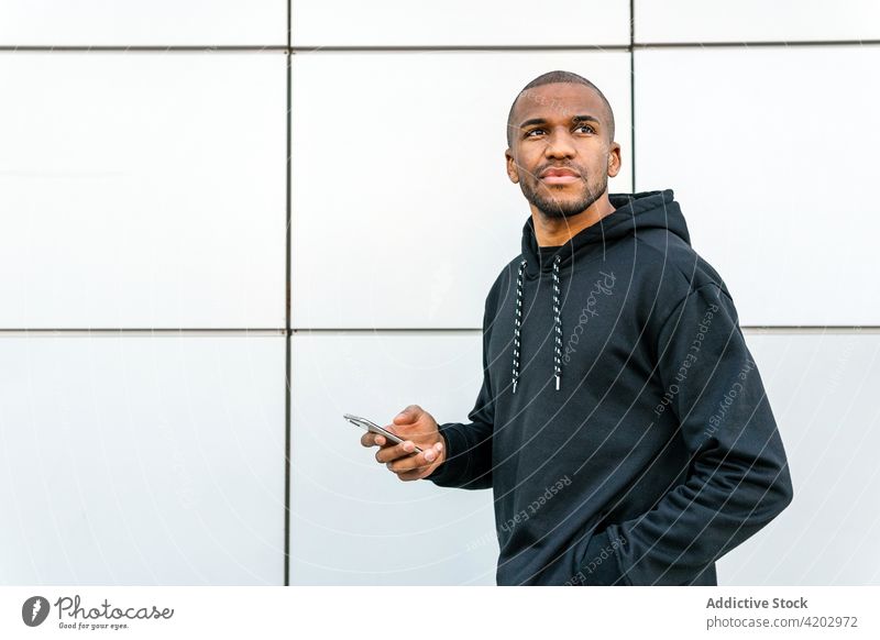 Masculine black man in hoodie near tiled wall hand in pocket style masculine smartphone browsing town city modern street contemplate message mobile phone