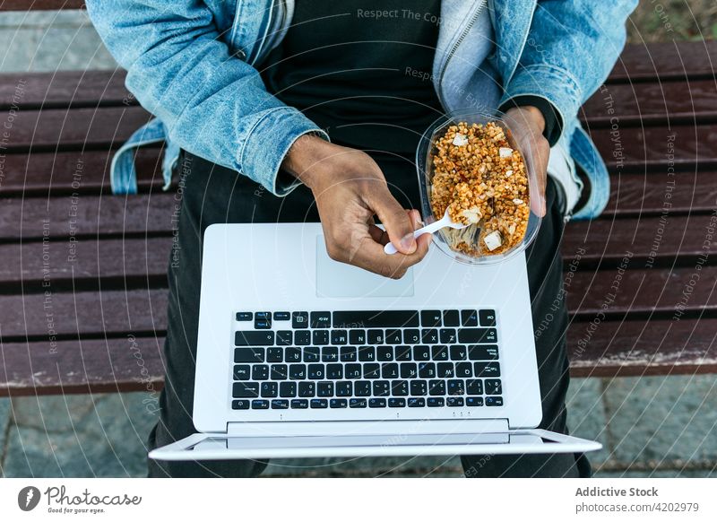 Crop black remote employee with laptop eating food on street lunch delicious dreamy man town worker freelance black screen contemplate denim jacket device